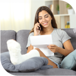 une femme assise sur son canapé, elle a une jambe dans le plâtre, elle est au téléphone souriante avec un papier à la main